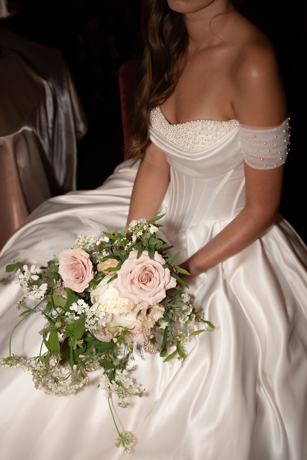 A bride in a strapless satin wedding dress with pearl embellishments on the bodice, holding a bouquet of pink and white roses and various white flowers, seated indoors.